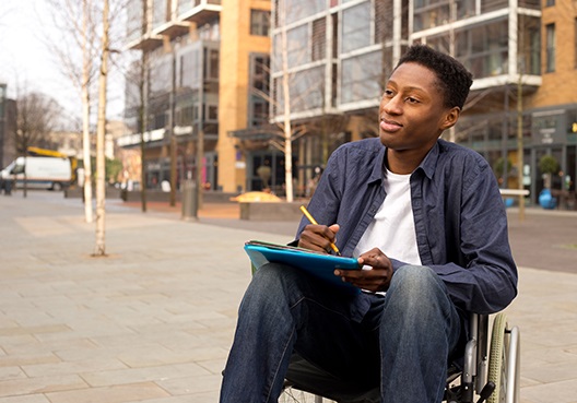A young man seated in a wheelchair writes down notes outdoors.