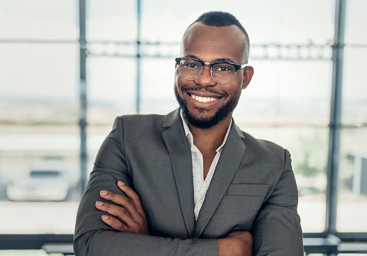 A man wearing a suit folds his arms and smiles at his place of work.