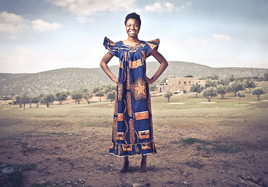 A woman wearing a blue and gold dress stands at the entrance of her village in Ghana.